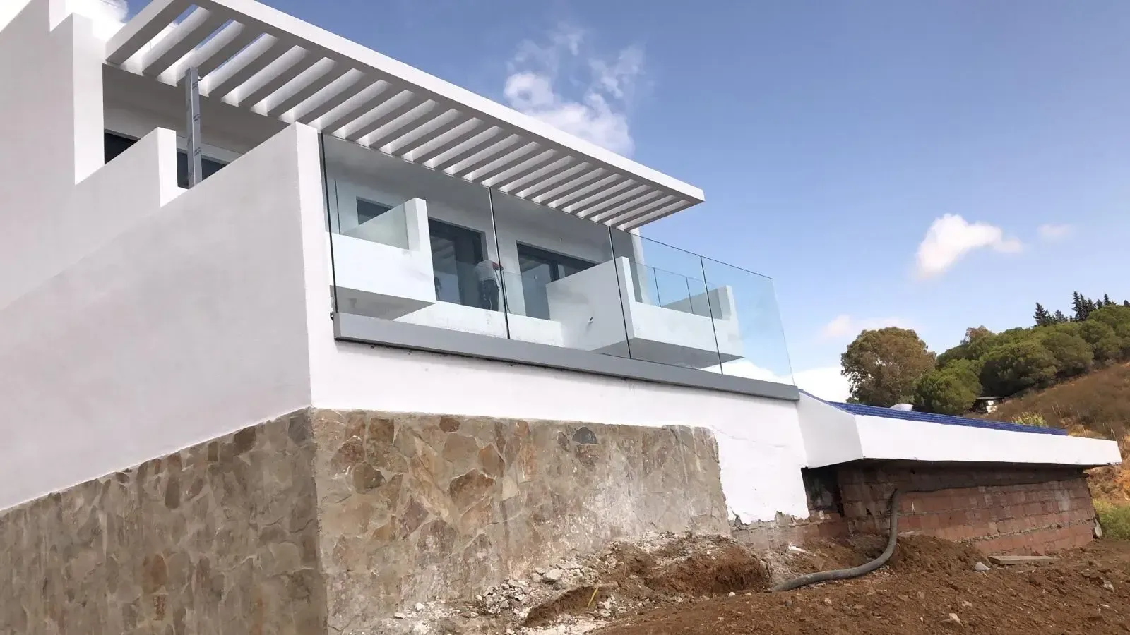 Una moderna casa blanca con barandillas de cristal en el balcón y cimientos de piedra, con un cielo azul de fondo y colinas al fondo.