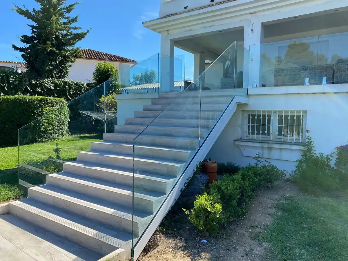 Escalera exterior de piedra con barandillas de cristal que conduce a una casa blanca con patio, situada en un jardín con árboles y arbustos.