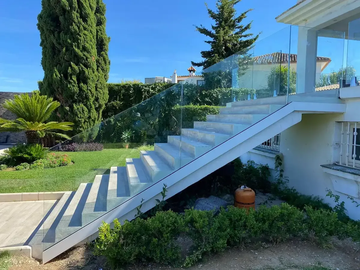 Escalera moderna de piedra con barandilla de cristal que conduce a un porche blanco, situado en un jardín soleado y bien cuidado.
