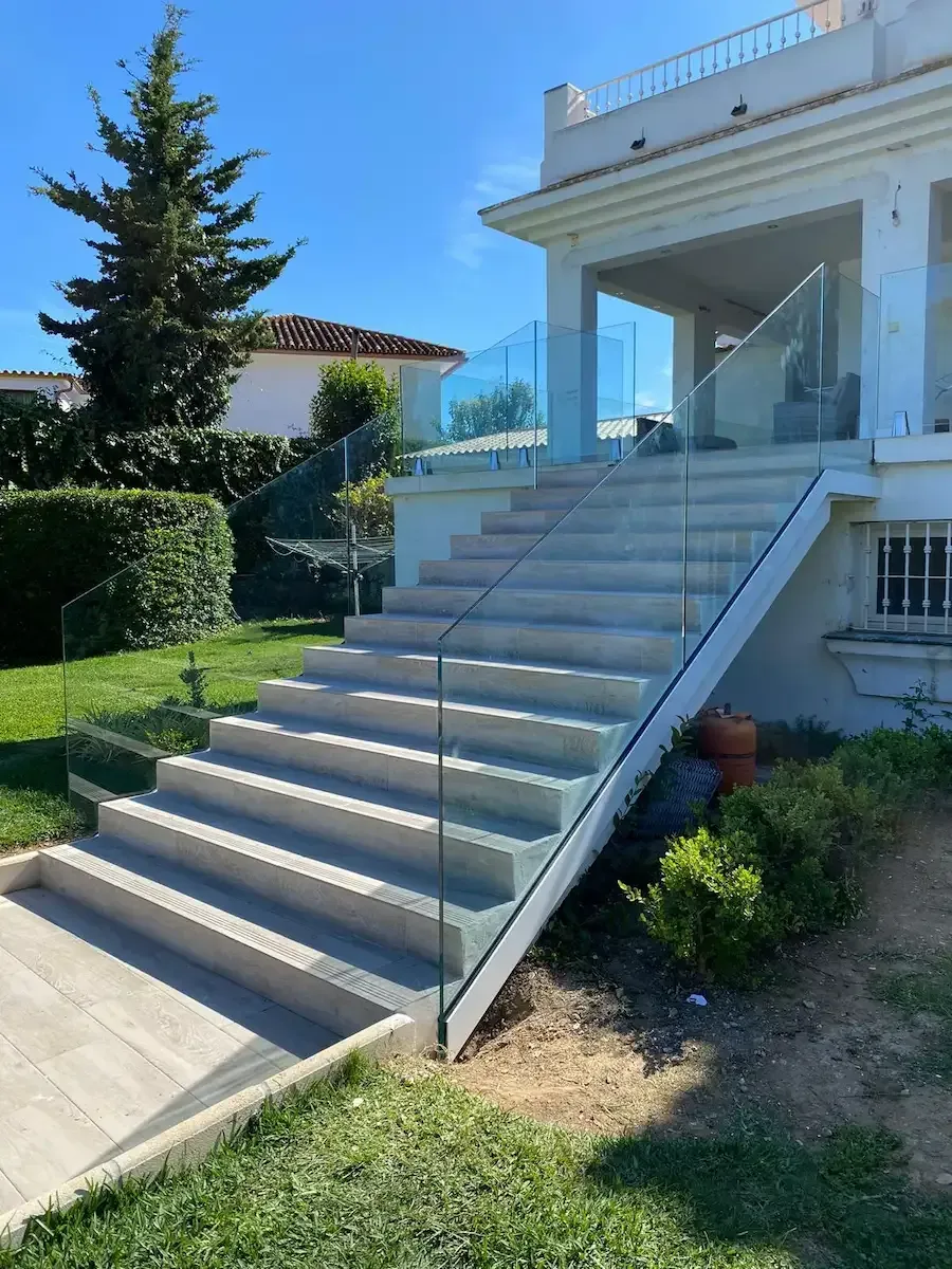 Escaleras de hormigón con una barandilla minimalista de cristal sin marco que conducen al patio de una casa blanca en un día soleado.