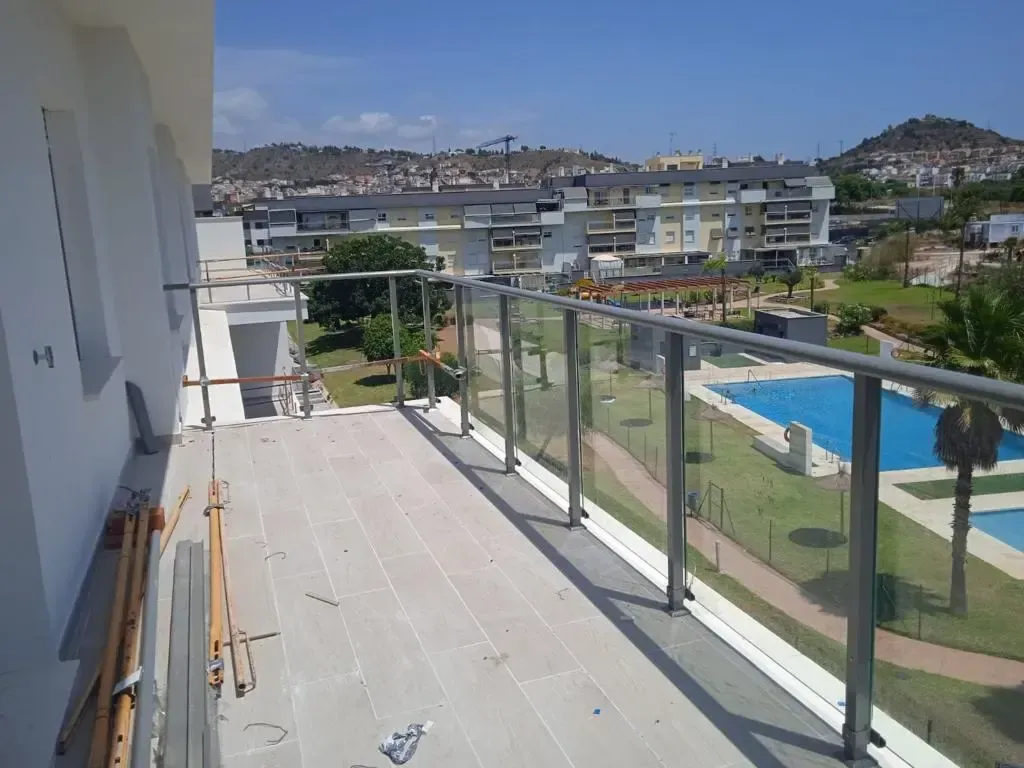 Un balcón con suelo de baldosas y barandilla de cristal ofrece vistas a una piscina y a edificios de apartamentos bajo un cielo azul despejado.