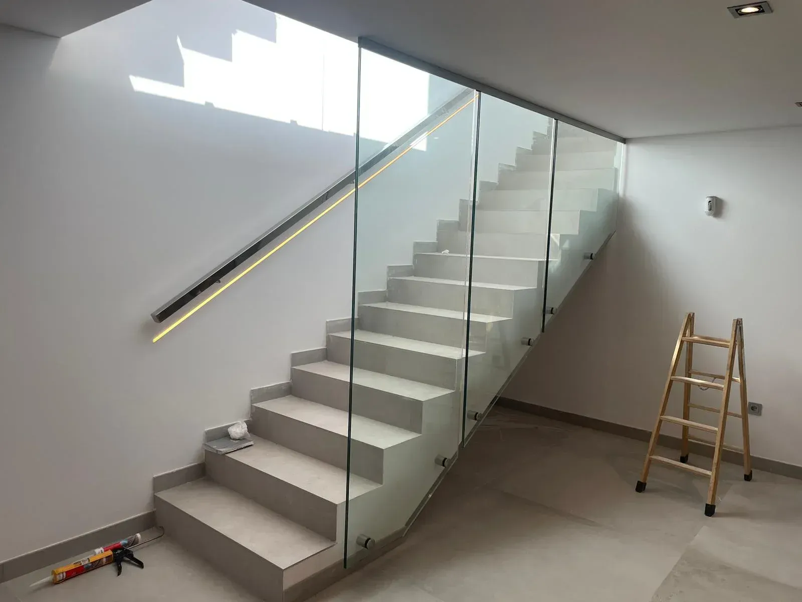 Una escalera minimalista con escalones de color claro, una barandilla de cristal moderna y un pasamanos mural con iluminación LED.