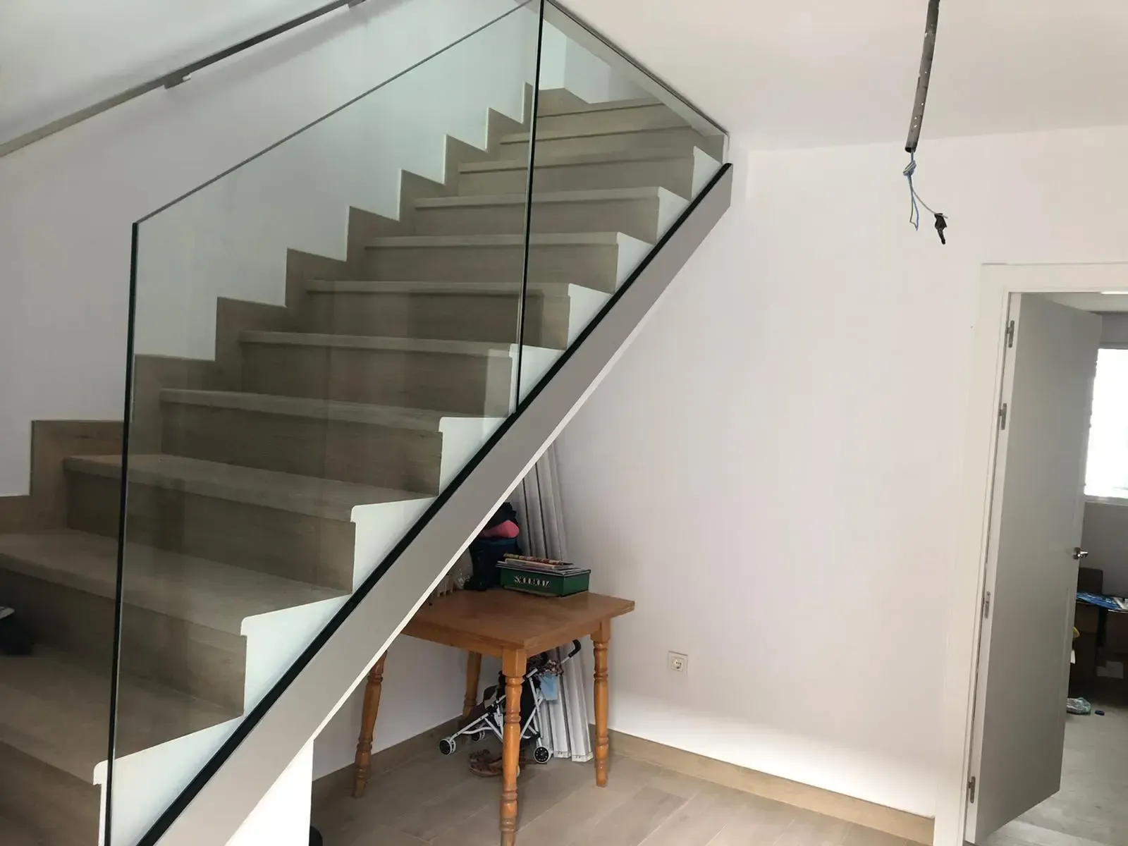 Escalera de madera clara con barandilla de cristal en una habitación moderna de paredes blancas con una pequeña mesa auxiliar de madera debajo.
