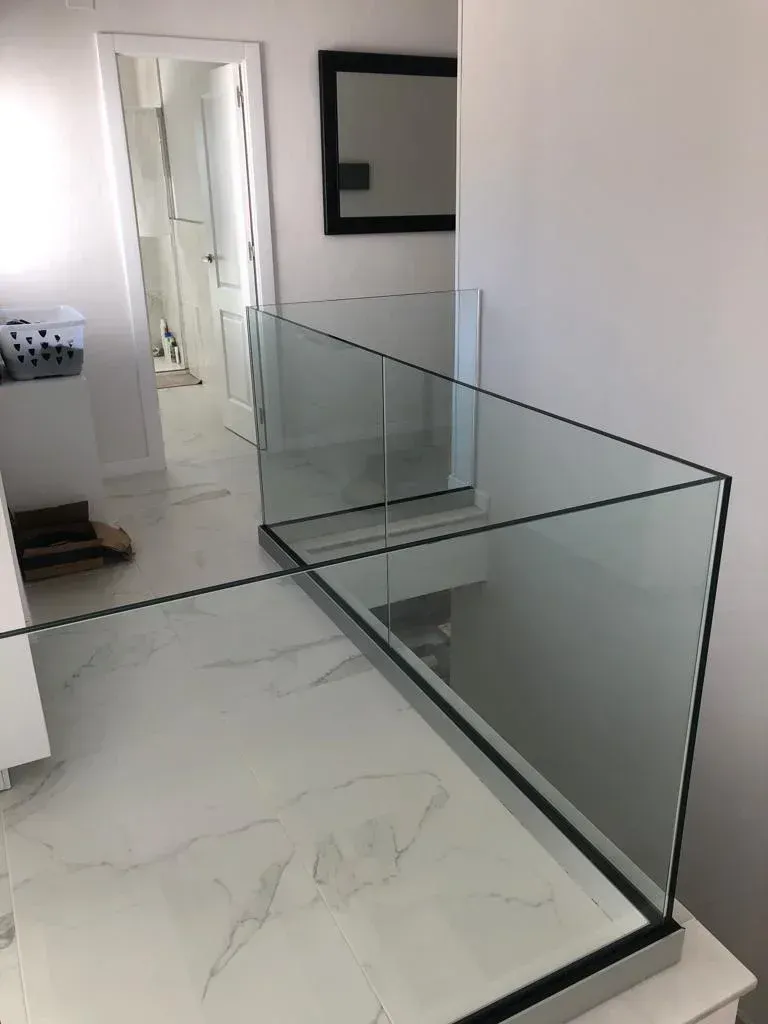 Barandilla de cristal sin marco sobre un suelo de mármol blanco, que bordea un rellano de escalera con una puerta al fondo.