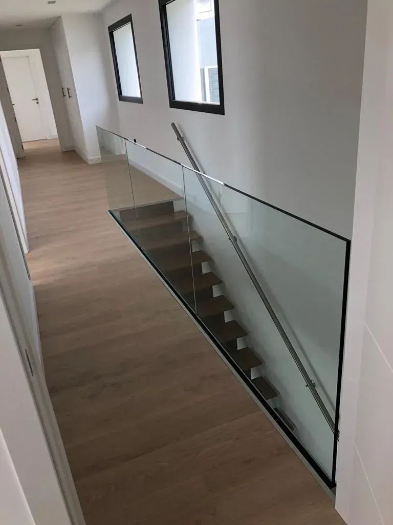 Un moderno recibidor interior con suelo de madera clara, que cuenta con una escalera con peldaños de madera y una barandilla de cristal sin marco.