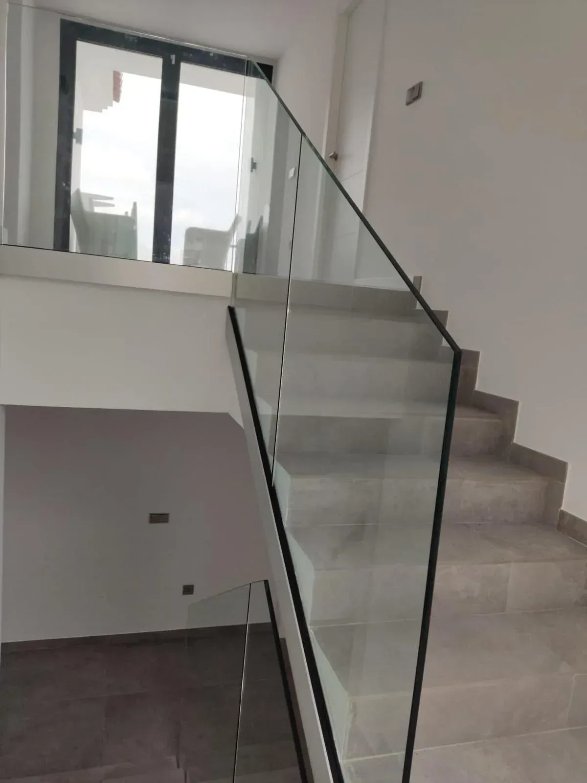 Escalera con peldaños de piedra gris y barandilla de cristal, que conduce a un rellano superior con una gran puerta de cristal.