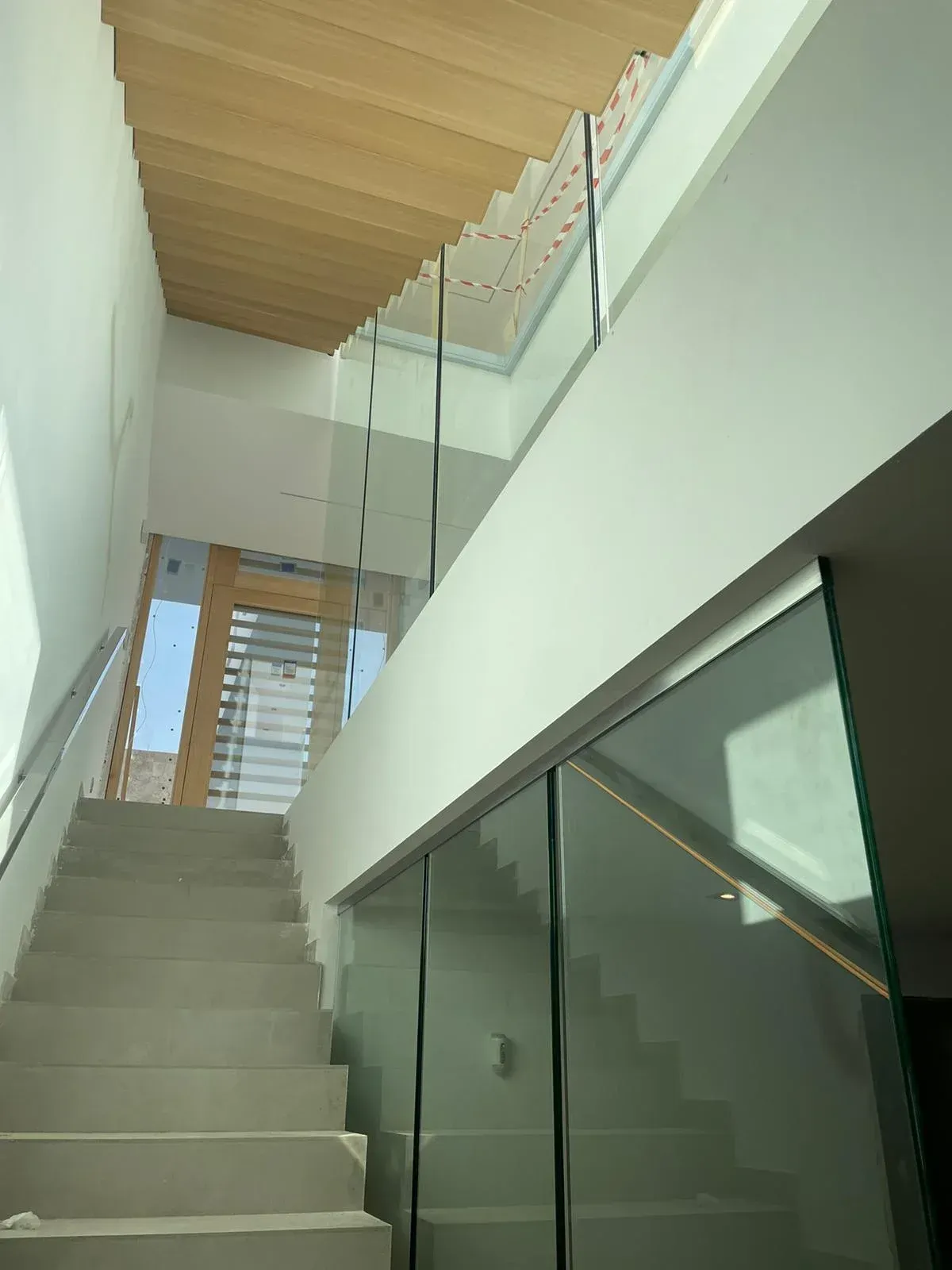Vista desde la parte inferior de una escalera de hormigón que conduce hacia arriba, con barandilla de cristal y techo revestido de paneles de madera.