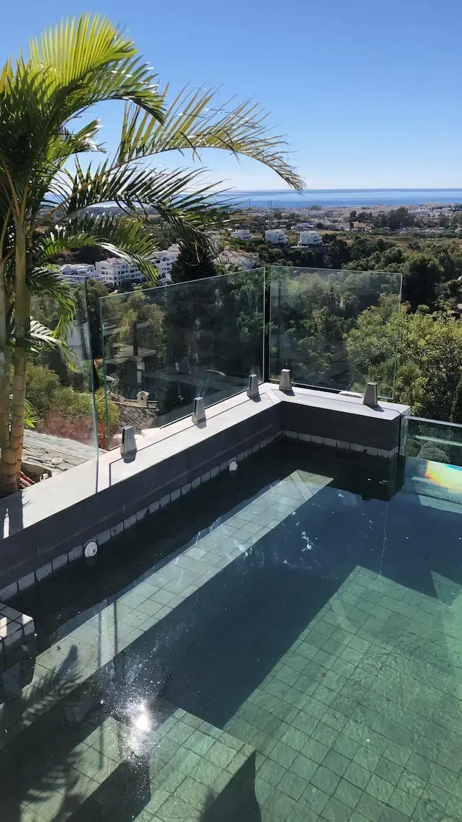 Una vista panorámica de una piscina infinita con azulejos oscuros que da a un paisaje soleado con vegetación y una vista lejana del mar.