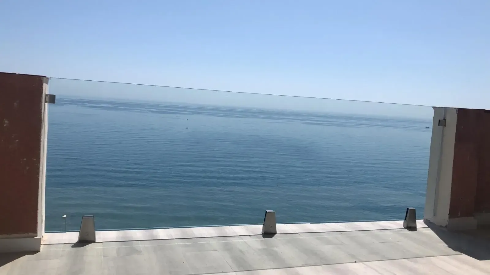 Vista de un mar azul desde un balcón con barandilla de cristal moderna y suelo de piedra bajo un cielo despejado y luminoso.