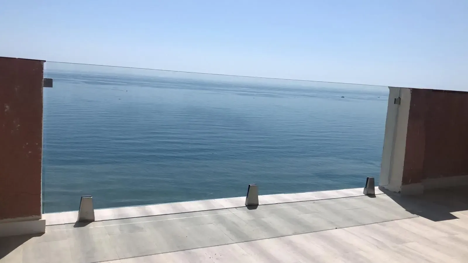 Una vista del océano desde un balcón con paredes rojas y barandilla de cristal bajo un cielo azul despejado.