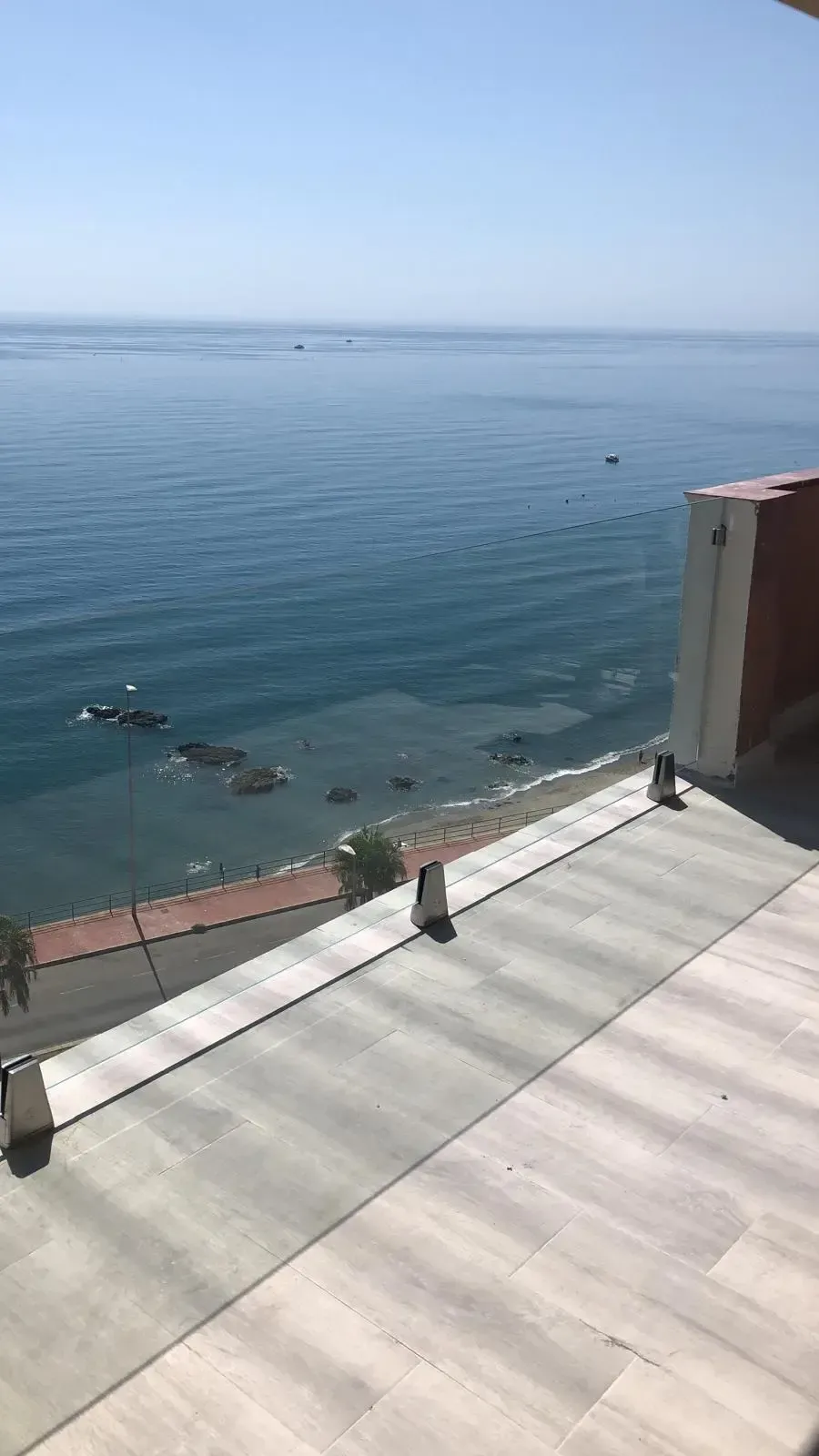 Una vista panorámica desde un balcón con vistas a un mar azul brillante, una costa rocosa y un paseo pavimentado debajo.