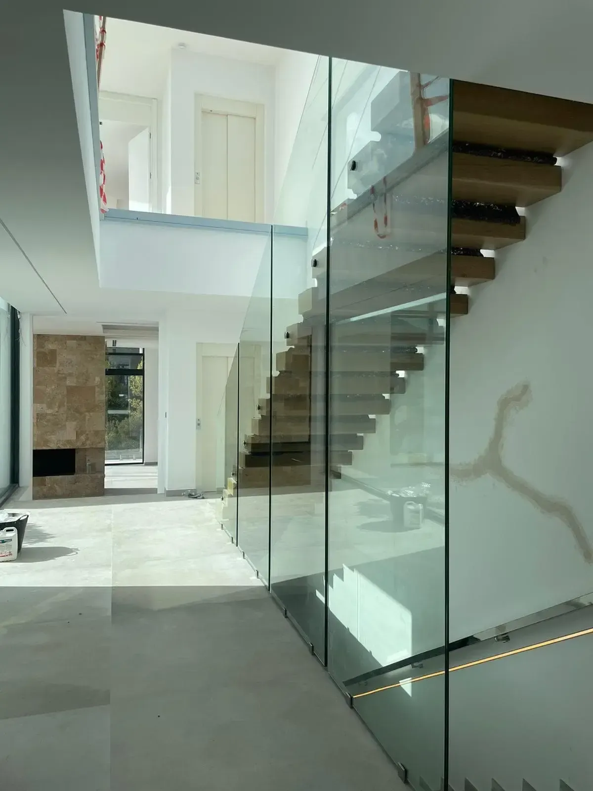 En una vivienda luminosa, el interior de estilo moderno presenta una escalera de madera flotante rodeada de altos paneles de vidrio, que contrasta con una pared blanca.