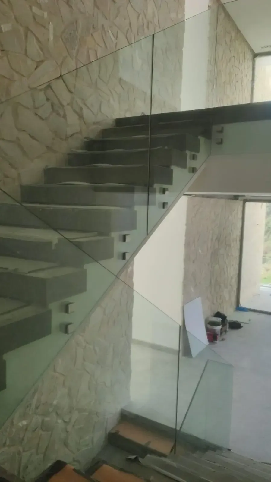 Escalera moderna con peldaños flotantes de hormigón y barandilla de cristal sin marco montada sobre una pared de piedra texturizada.