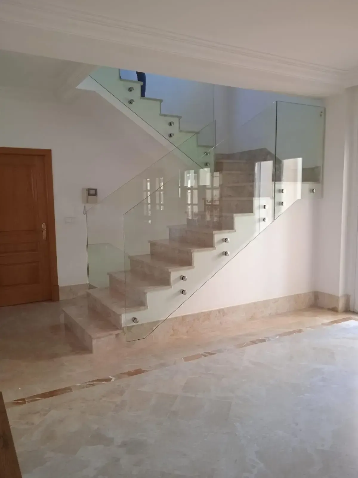 Una escalera con escalones de piedra beige y una moderna barandilla de cristal sin marco con separadores metálicos en un pasillo alicatado.