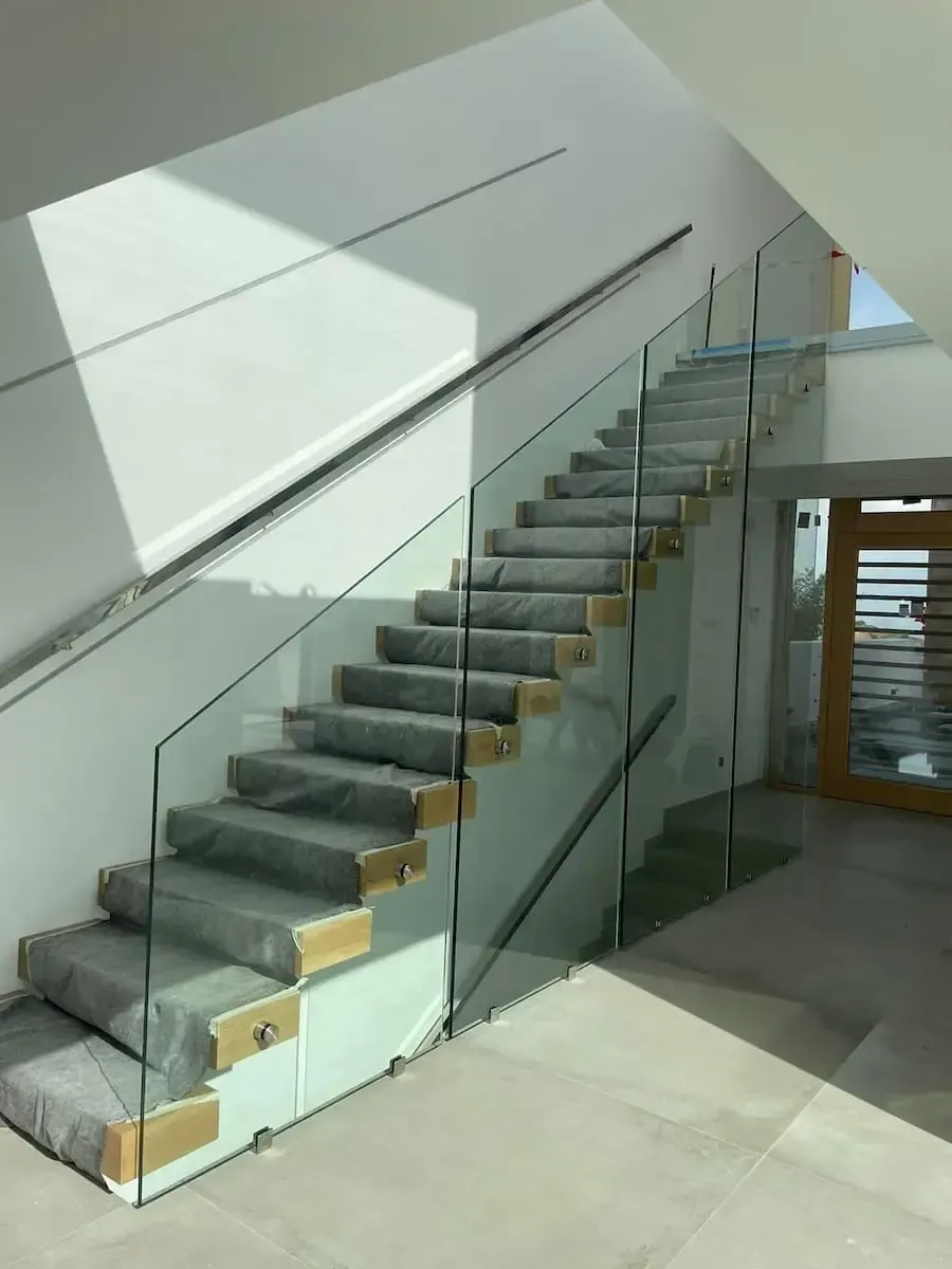 Escalera moderna con escalones alfombrados en gris, detalles de madera y una barandilla de cristal transparente con pasamanos de metal.