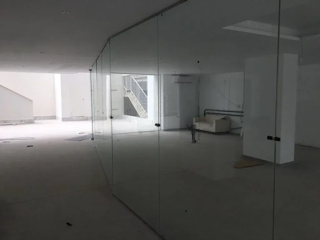 Una gran mampara de cristal que va desde el suelo hasta el techo separa una habitación blanca vacía de una zona que cuenta con un sofá y una escalera.