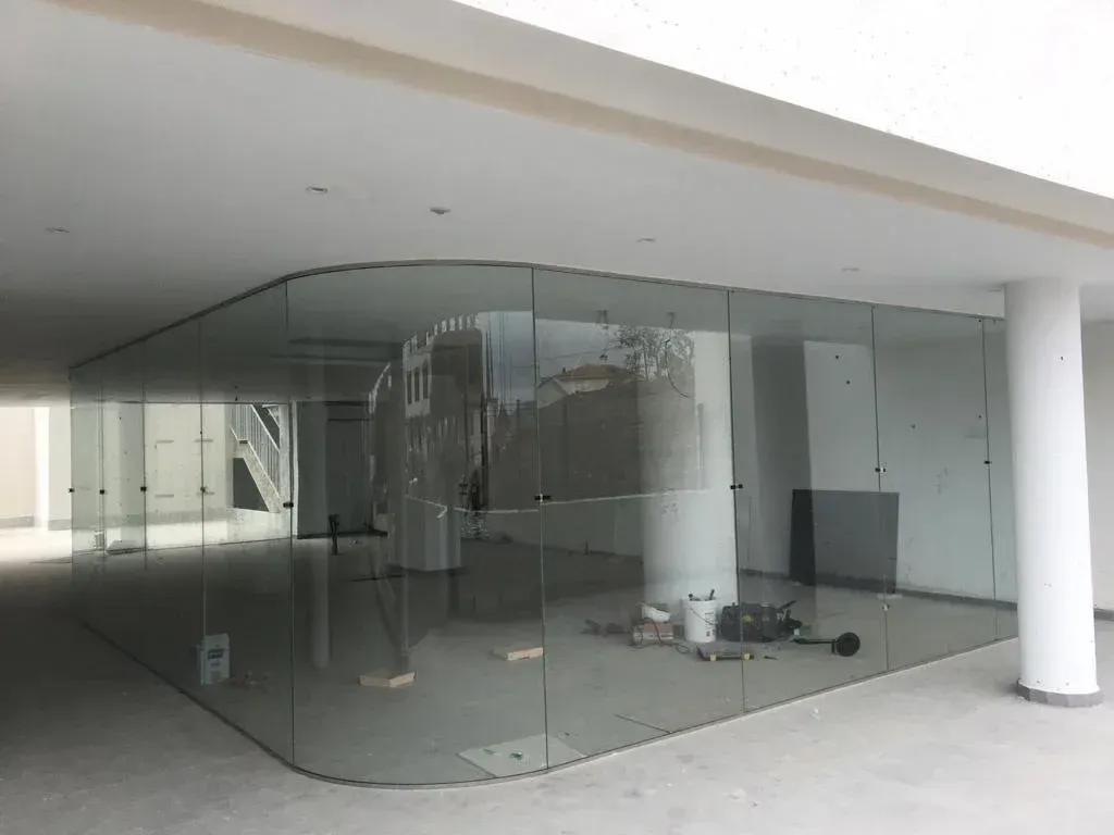 Paredes curvas de vidrio dividen un espacio comercial moderno y vacío bajo un toldo blanco, con herramientas de construcción en su interior.