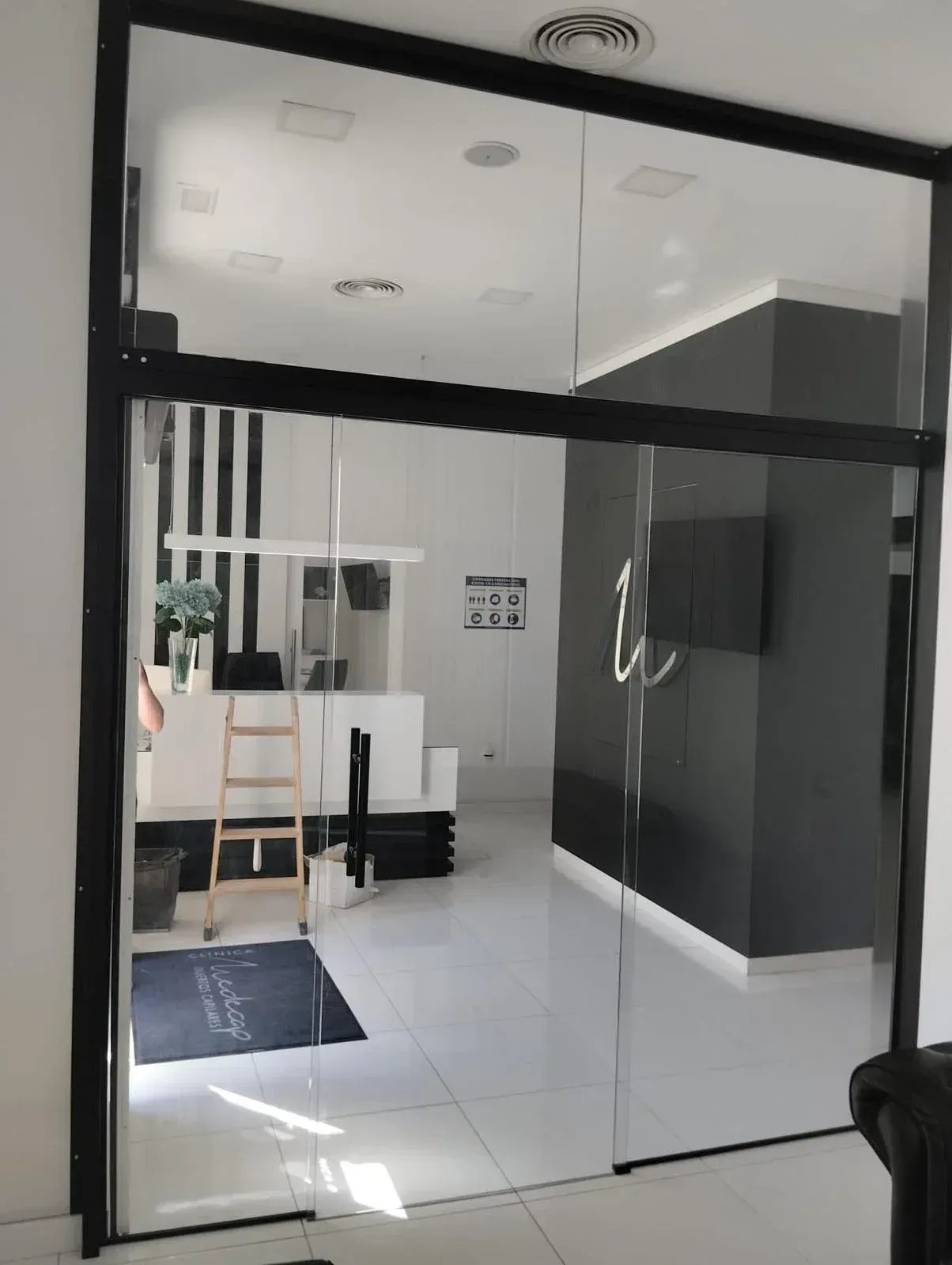 Se puede ver, a través de una puerta de cristal, una oficina moderna con paredes blancas, una pared decorativa negra, una escalera y un mostrador de recepción.