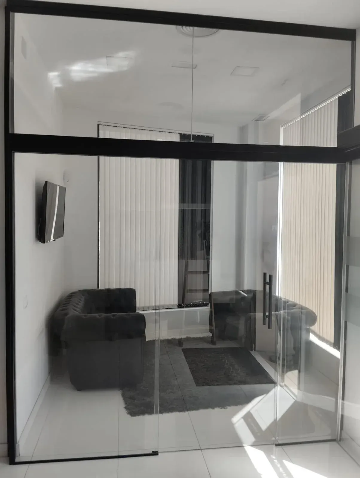 Vista a través de una mampara de cristal de una oficina hacia un salón moderno con un sofá oscuro y una alfombra en una habitación luminosa de paredes blancas.