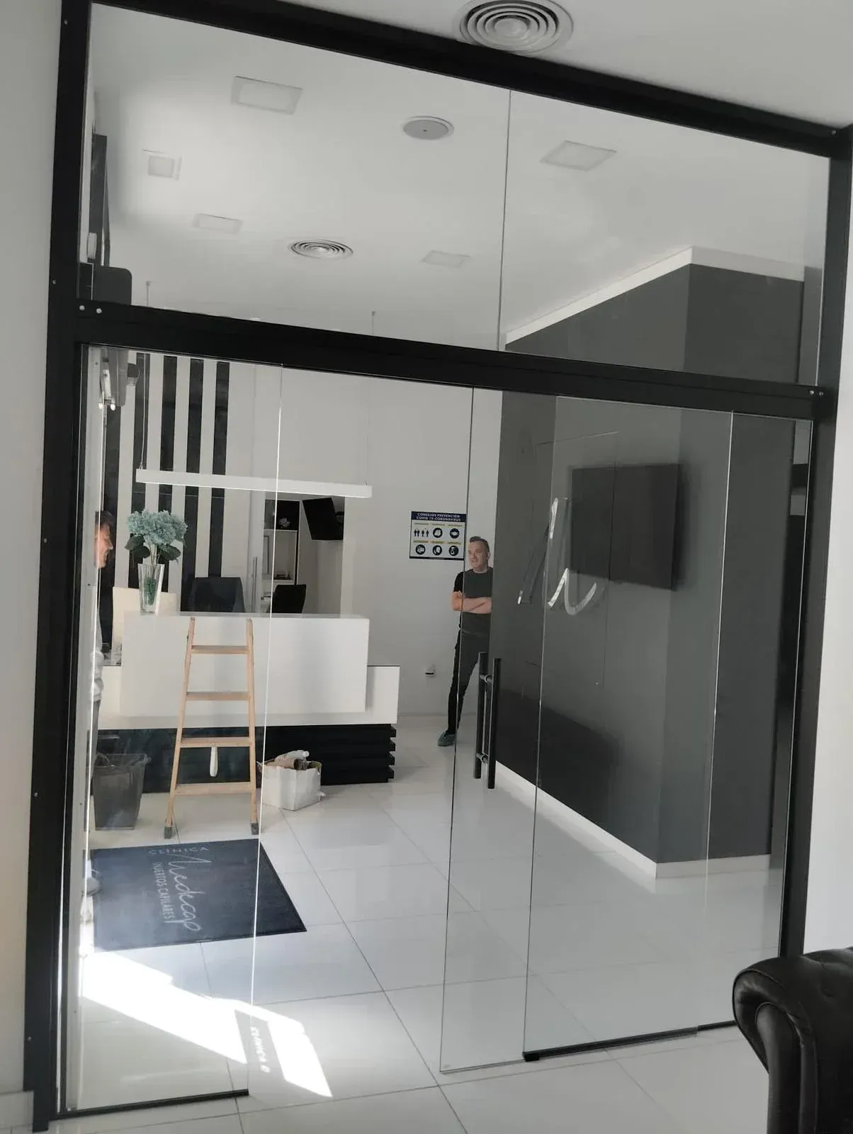 Una vista a través de una puerta de cristal con marco negro hacia una oficina moderna con suelos blancos, una escalera de madera y una pared decorativa gris.