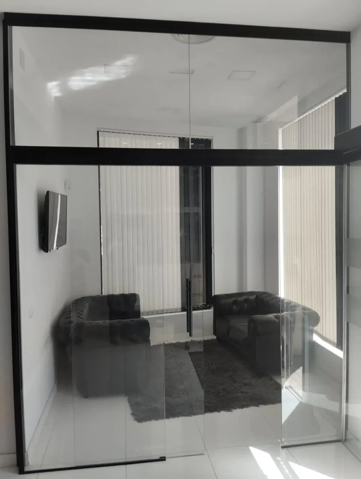 Un pequeño espacio de oficina rodeado de paredes de cristal, con dos sillas de cuero negro, una alfombra oscura y un televisor montado en la pared.