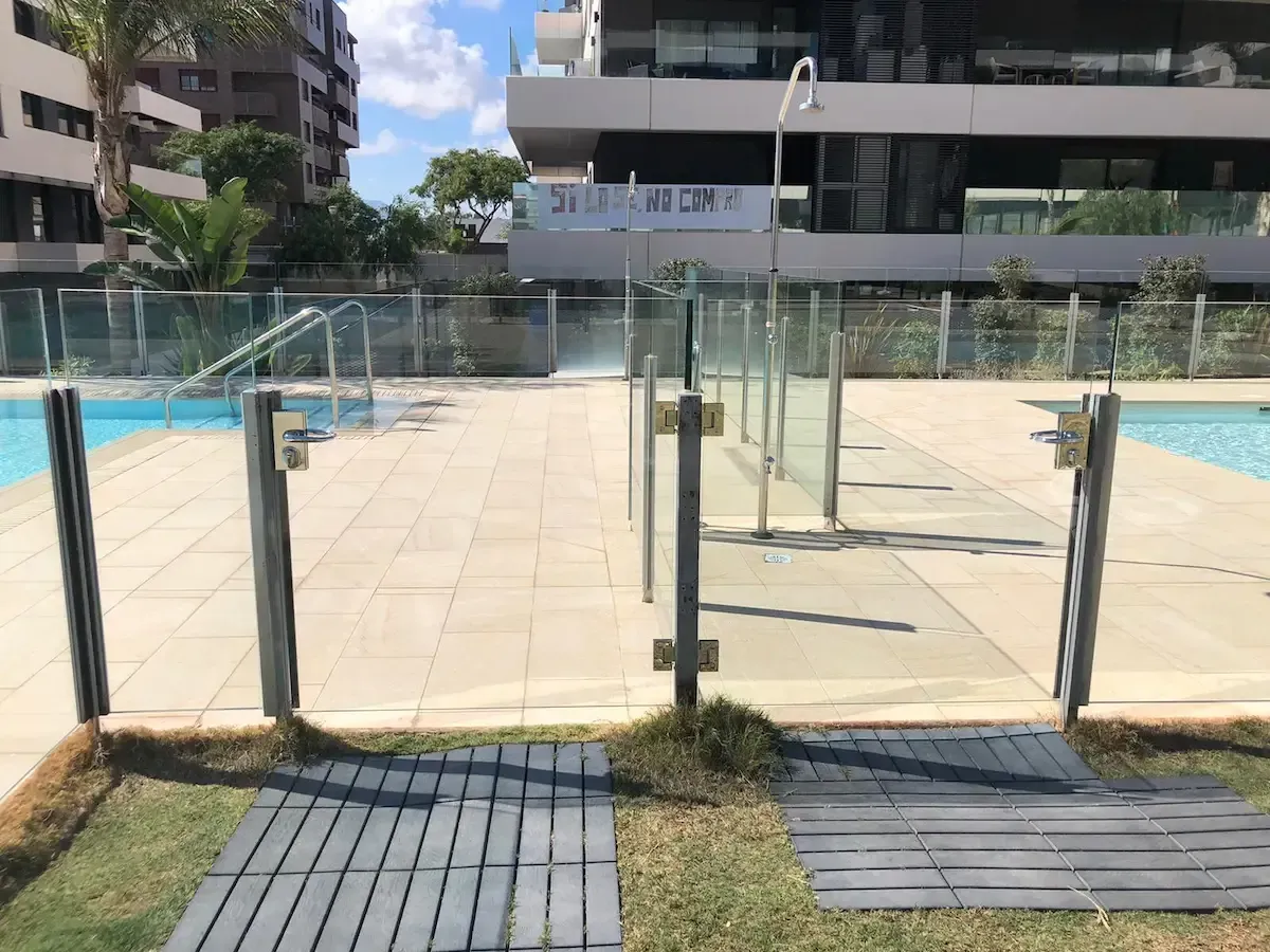 Dos entradas cerradas con paneles de vidrio dan acceso a una terraza pavimentada junto a la piscina, que cuenta con ducha exterior y tumbonas.