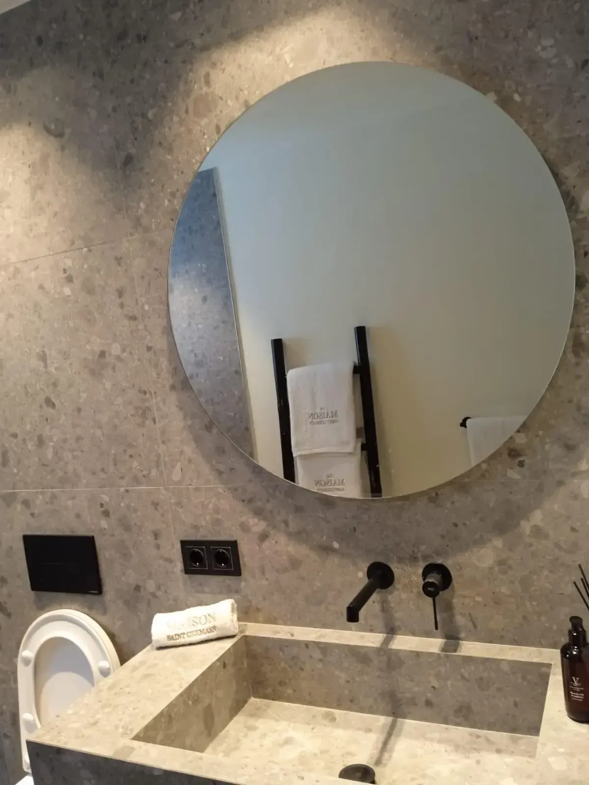 Un moderno mueble de baño con lavabo cuadrado de piedra, grifo negro montado en la pared y espejo redondo sobre azulejos moteados.