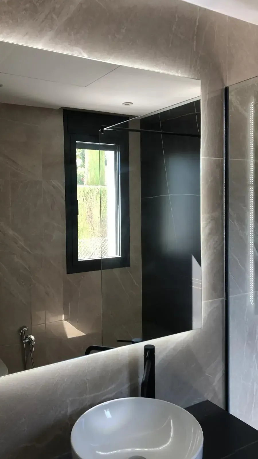 Baño moderno con lavabo redondo blanco sobre encimera, grifo negro y un gran espejo de pared retroiluminado que refleja una ventana.