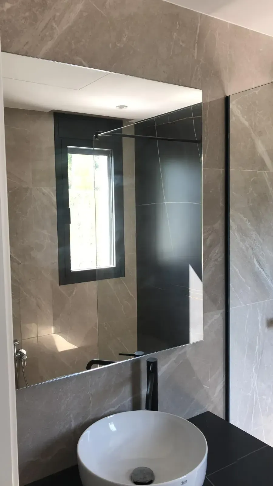 Un baño moderno con lavabo blanco sobre un mueble oscuro, un gran espejo rectangular y paredes alicatadas en gris.