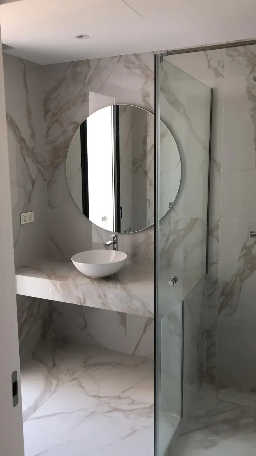 Baño moderno con azulejos de mármol blanco, lavabo suspendido, un gran espejo redondo y una ducha con mampara de cristal.