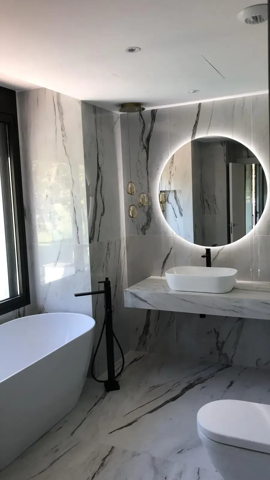 Un baño moderno con bañera de mármol, tocador flotante de mármol con lavabo sobre encimera y espejo circular con iluminación LED.