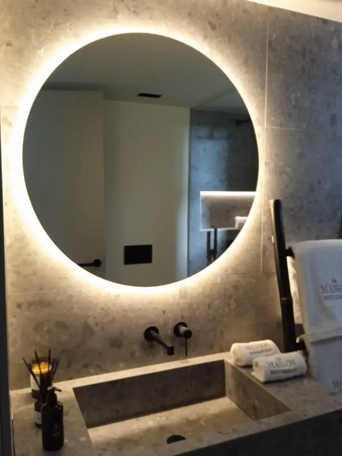 Un espejo circular retroiluminado cuelga sobre un tocador de baño de piedra con un lavabo cuadrado y grifería negra montada en la pared.