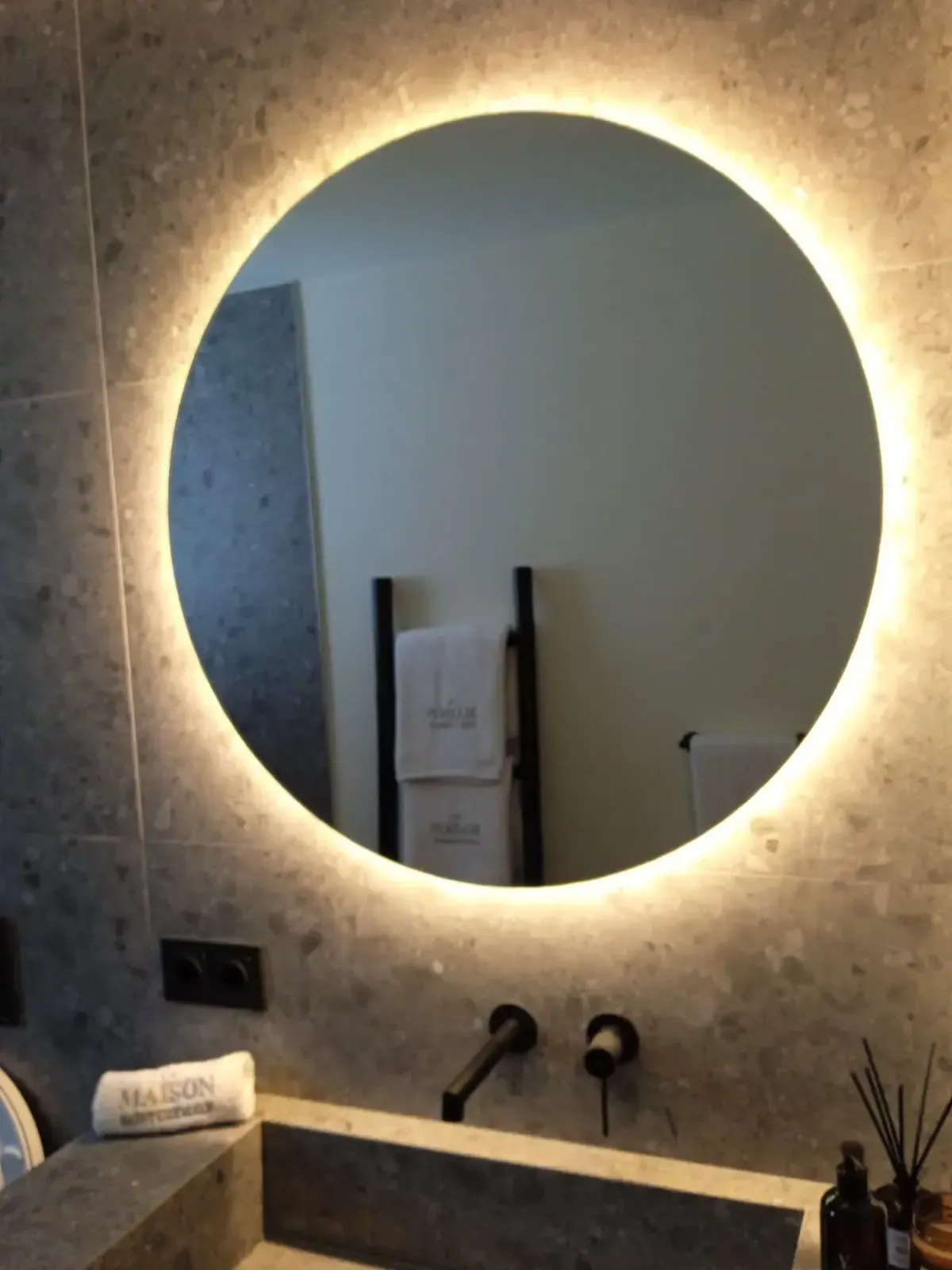 Un espejo circular retroiluminado cuelga sobre un tocador de piedra con un grifo moderno de color negro mate, frente a una pared de azulejos grises texturizados.