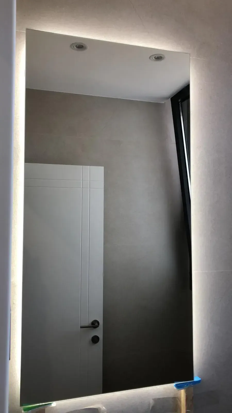 Un espejo rectangular con paneles LED integrados con iluminación lateral, montado sobre una pared gris, refleja una puerta blanca cerrada.