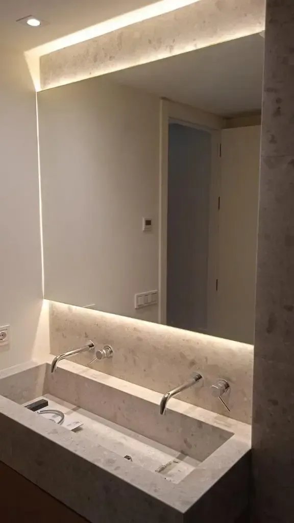 Lavabo doble moderno de piedra con grifería mural y un gran espejo rectangular retroiluminado en un baño contemporáneo.