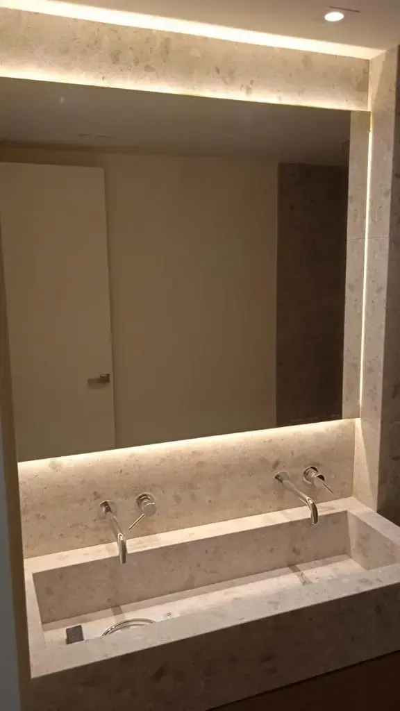 Un moderno tocador con doble lavabo, fabricado en piedra beige, con grifería mural e iluminación LED empotrada.