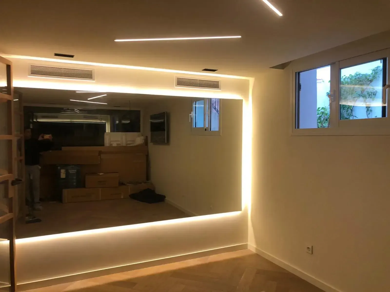Una habitación moderna con un gran espejo montado en la pared con retroiluminación LED integrada, suelos de madera noble y una pequeña ventana.