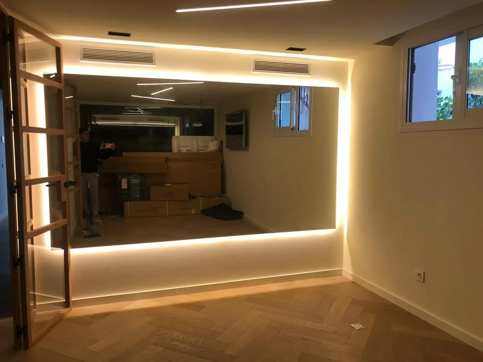 Un gran espejo de pared con retroiluminación LED integrada en una habitación con suelo de madera en espiga.