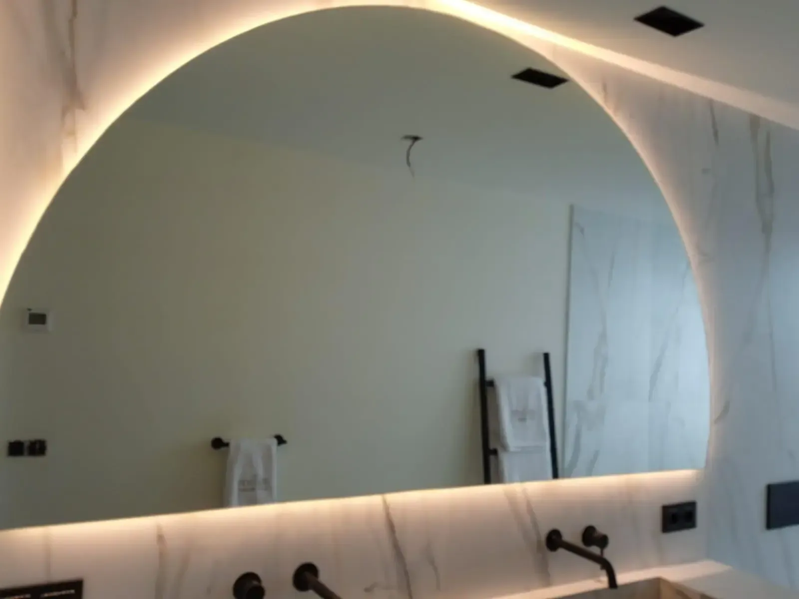 Un gran espejo de baño arqueado e iluminado desde atrás, montado sobre una pared de mármol blanco encima de un lavabo doble.