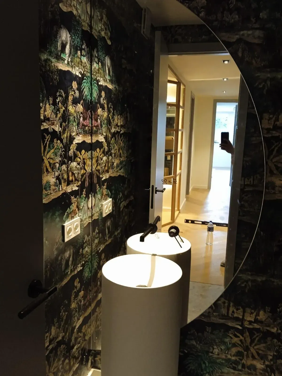 Dos lavabos cilíndricos blancos con pedestal se sitúan frente a una pared pintada con un papel pintado oscuro de intrincado diseño y un espejo circular.