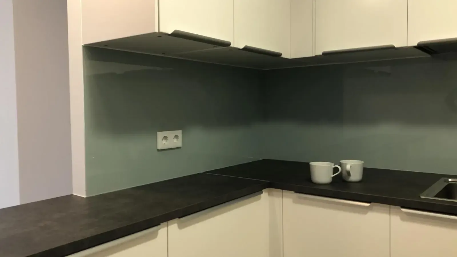 Un rincón de cocina con armarios blancos, una encimera oscura, un protector contra salpicaduras de vidrio verde salvia y dos tazas blancas.