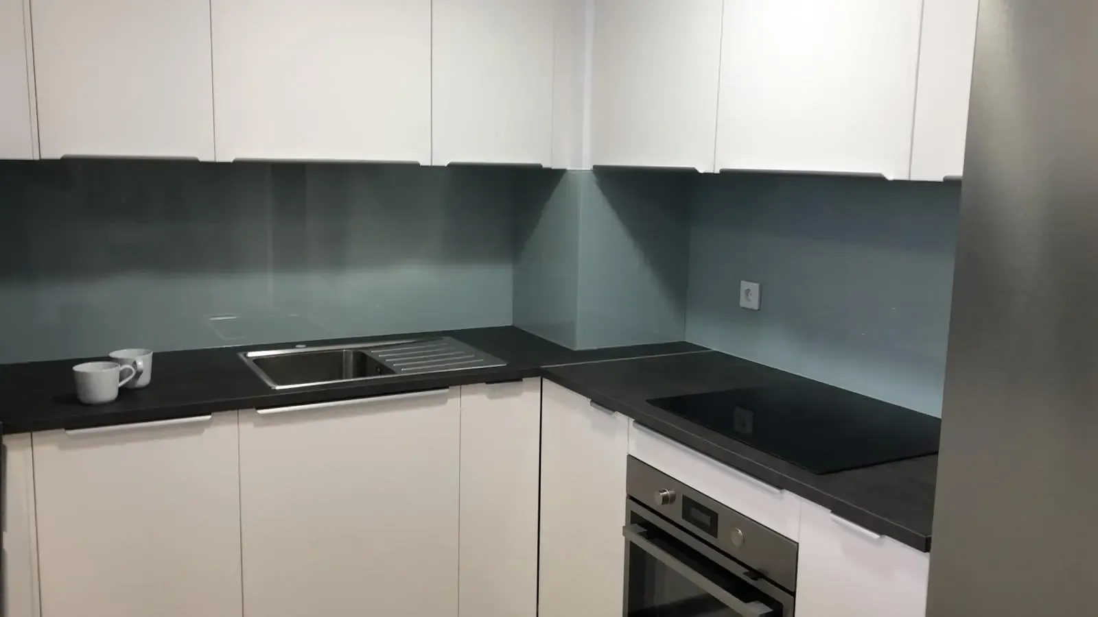 Una cocina moderna con armarios blancos, encimeras oscuras, fregadero de acero inoxidable y protector contra salpicaduras de vidrio azul grisáceo.