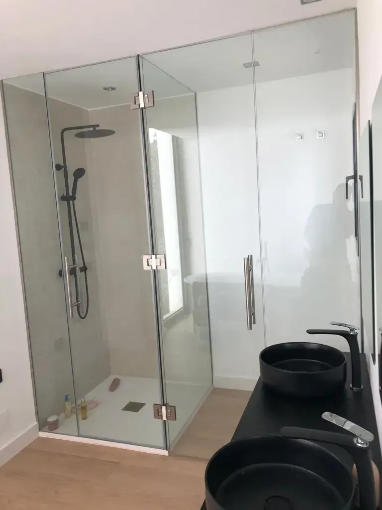 Un baño moderno con ducha acristalada, grifería negra y dos lavabos negros sobre un mueble de baño.