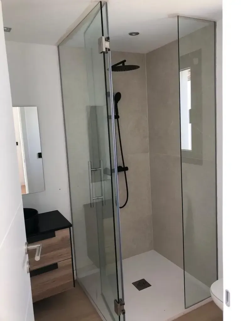 Un baño moderno con una ducha acristalada con grifería negra mate, junto a un tocador de madera con lavabo.
