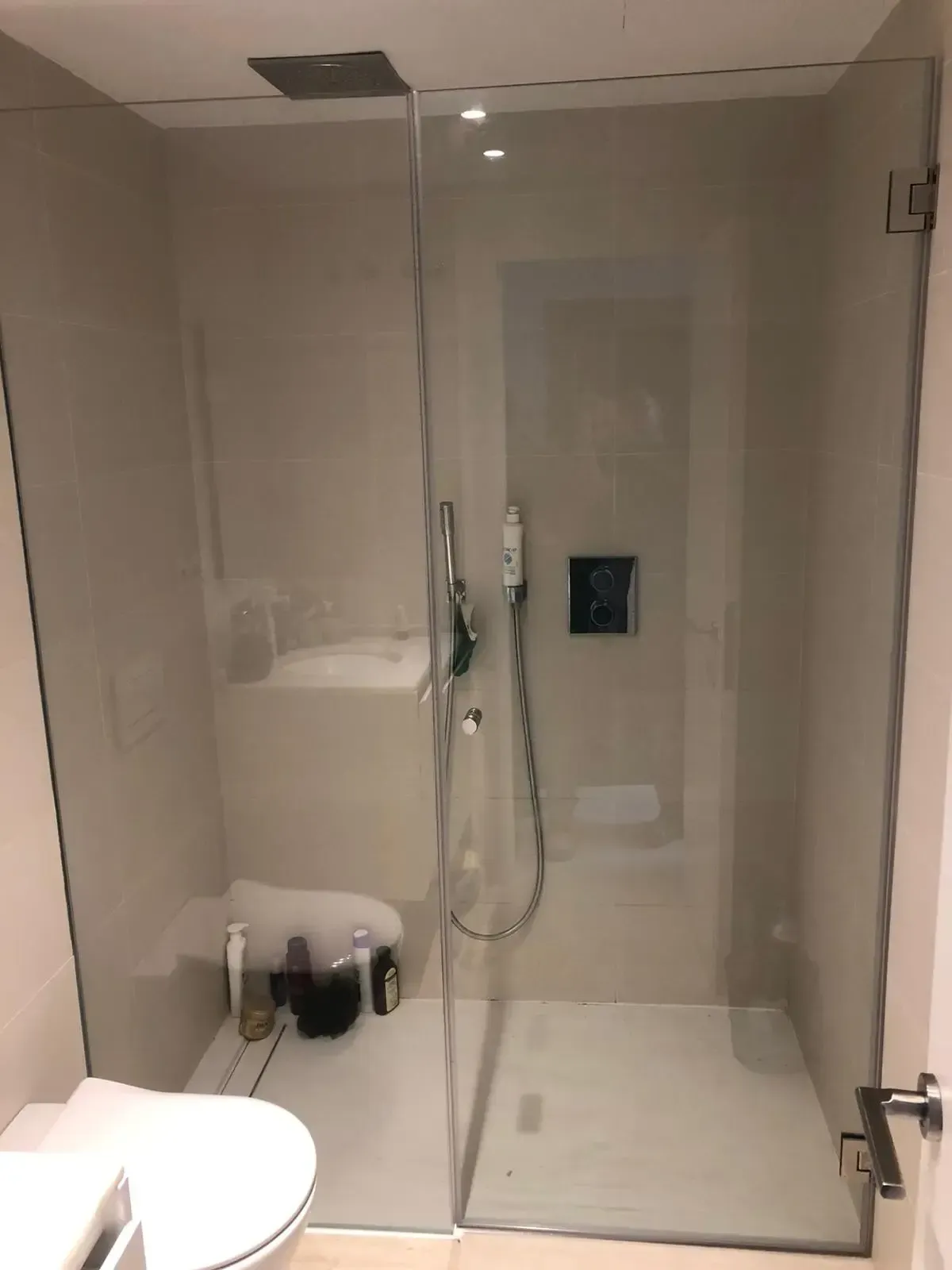 Una moderna cabina de ducha de cristal con paredes alicatadas en color beige, una ducha de mano y un cabezal de ducha tipo lluvia en un baño.