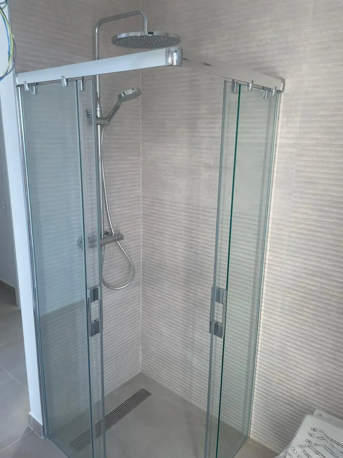 Una moderna cabina de ducha de esquina con puertas de cristal, un cabezal de ducha de efecto lluvia plateado y una ducha de mano, instalada sobre paredes alicatadas.