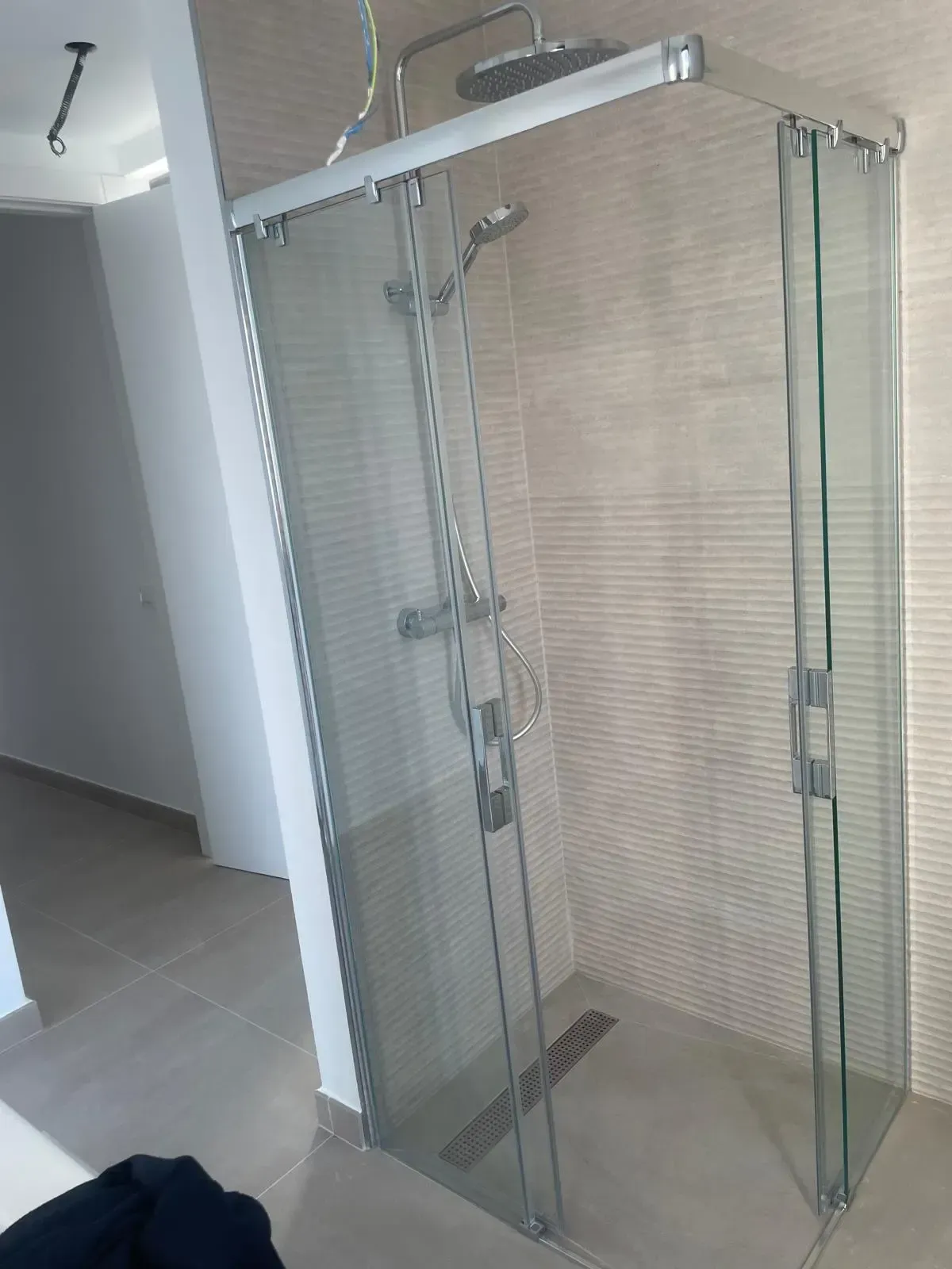 Una cabina de ducha acristalada con cabezal de ducha tipo lluvia y rociador de mano, con azulejos de pared texturizados de color claro.