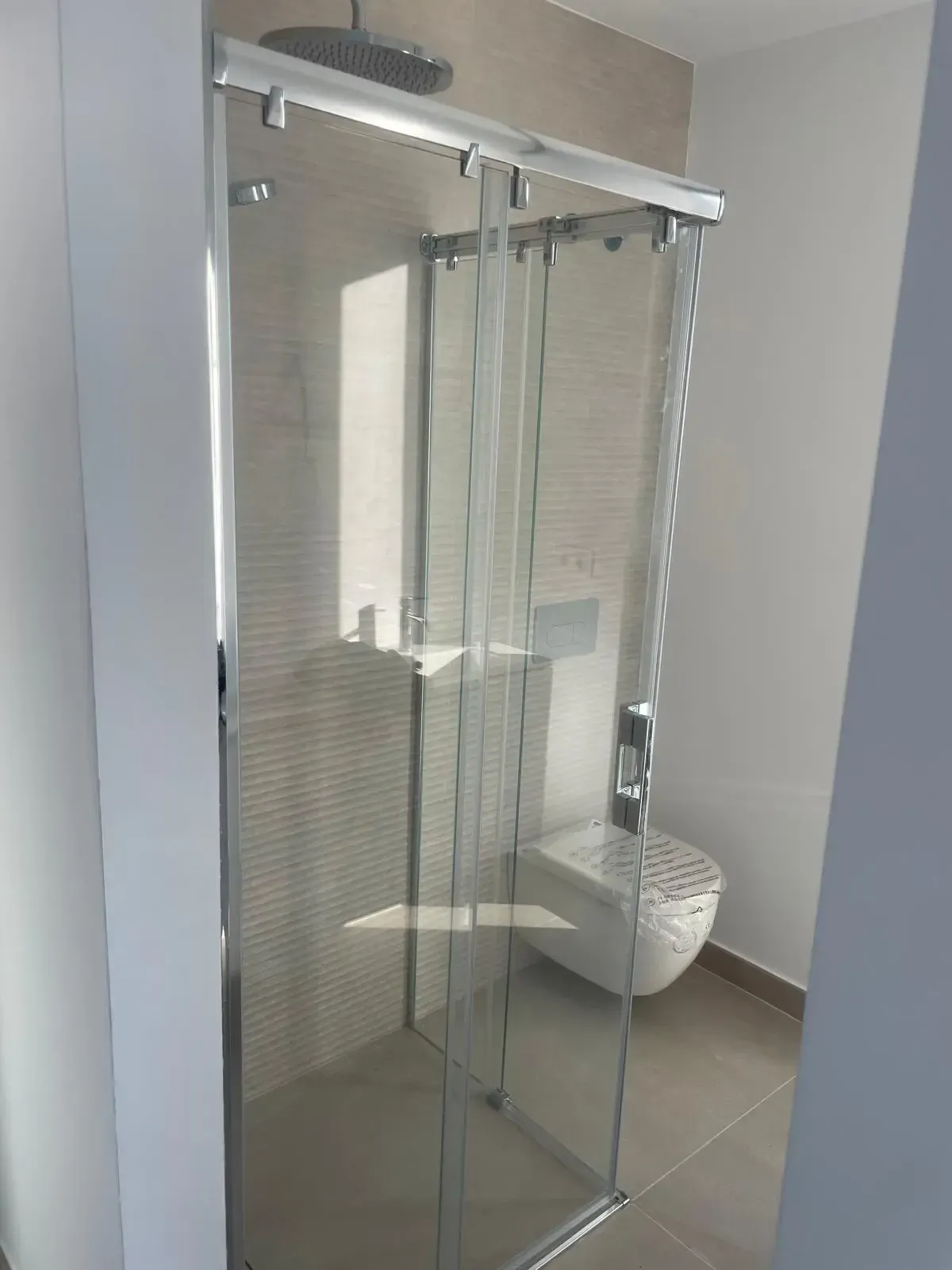 Una moderna cabina de ducha de cristal con puertas correderas y cabezal de ducha tipo lluvia, situada en un baño alicatado en tonos claros.