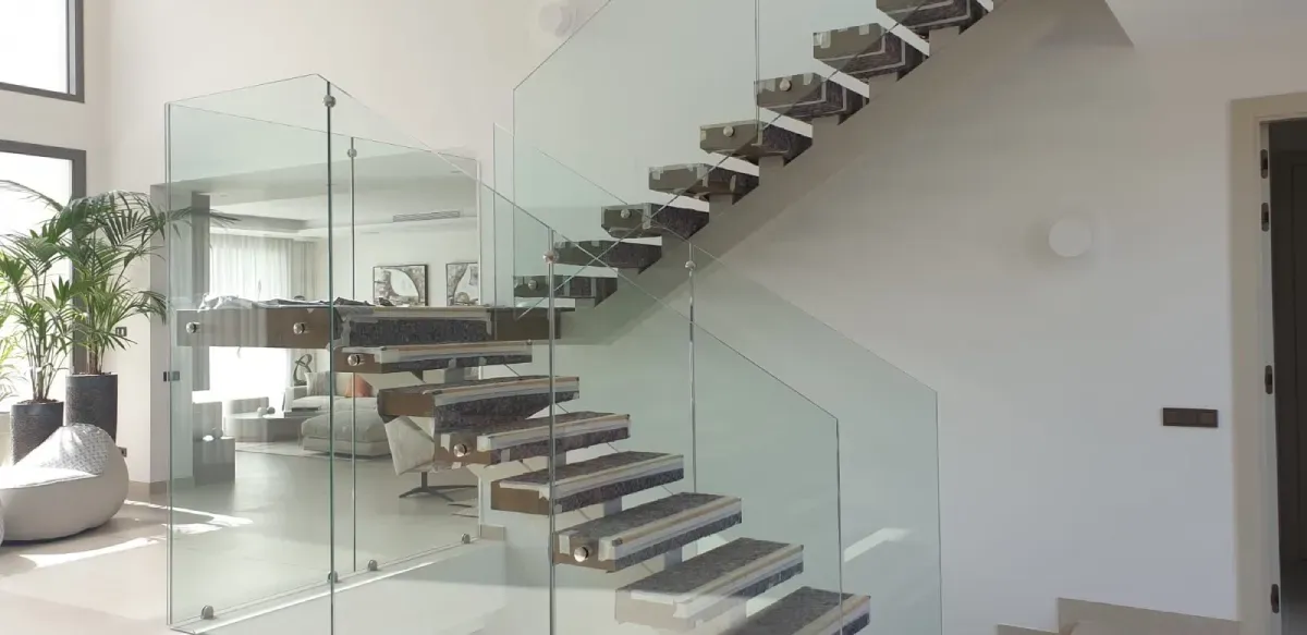 Una escalera con peldaños de piedra oscura y barandilla de cristal transparente en un interior luminoso y minimalista.