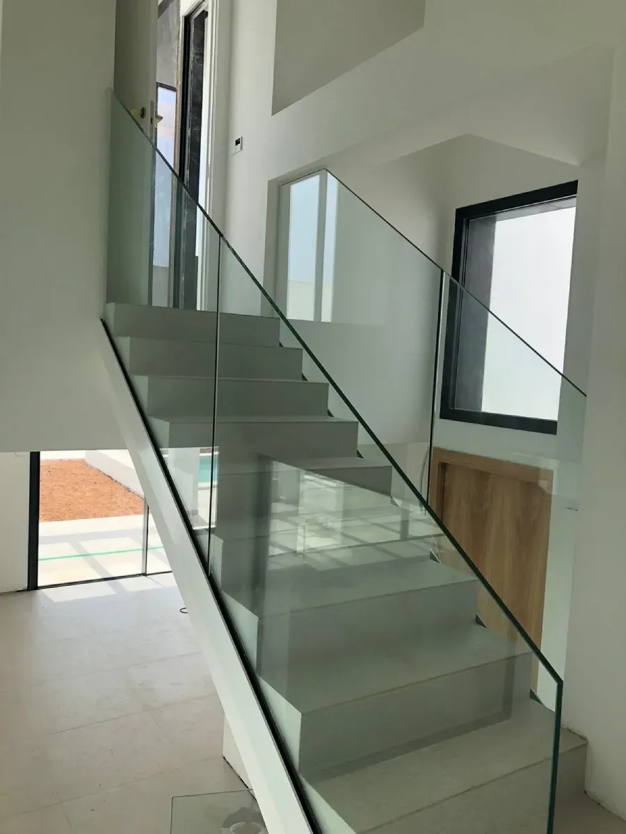 Una moderna escalera interior con escalones blancos y barandilla de cristal, ubicada en una casa luminosa y minimalista.